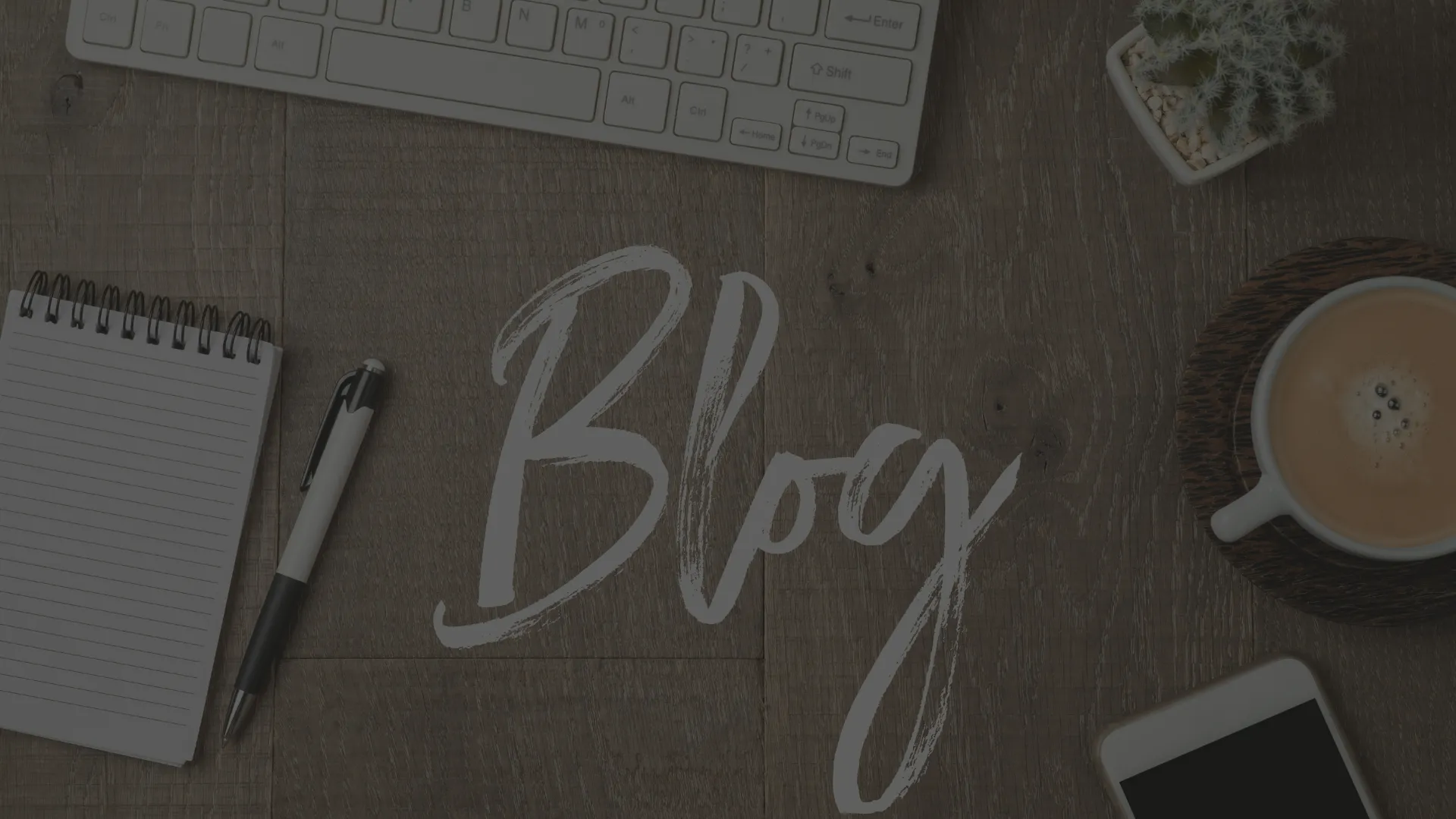 Un plan de travail en bois sur lequel est posé un clavier d'ordinateur, une plante, un café, un téléphone, un bloc-notes et le mot "Blog" écrit au milieu.